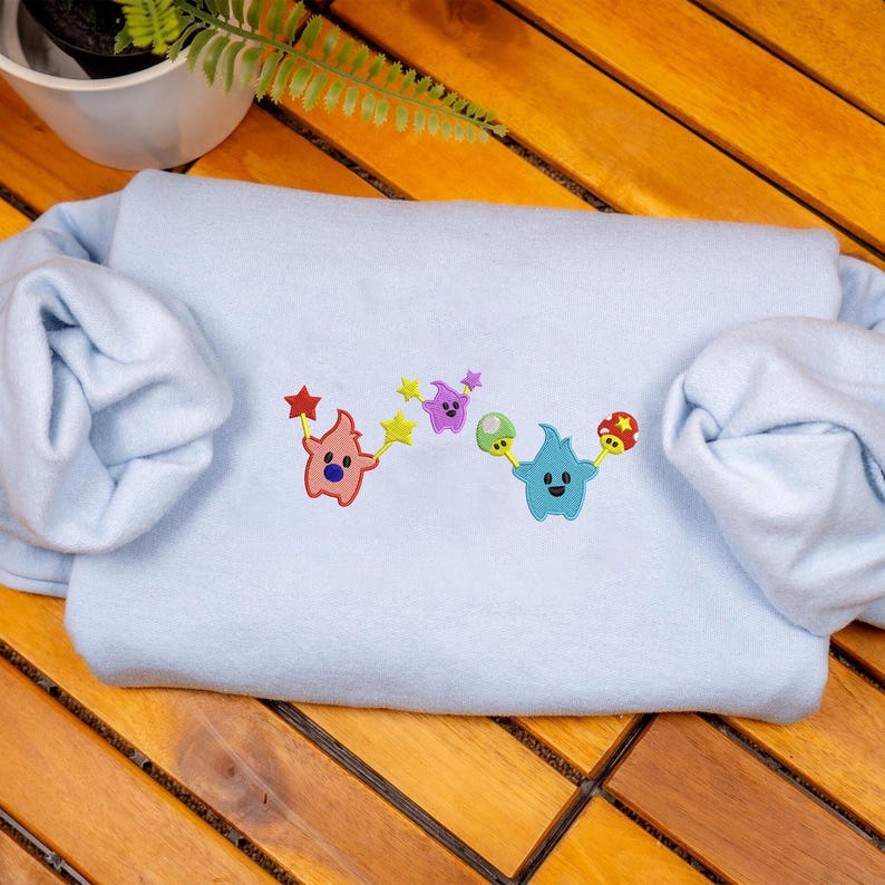 May include: A light blue sweatshirt with embroidered cartoon characters. The design features a pink character holding a red star, a purple character, a green character, and a blue character holding a mushroom.