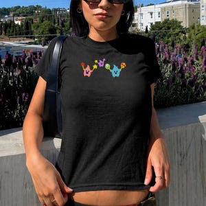 May include: Black crew neck t-shirt with colorful cartoon characters embroidered on the front. The design features two whimsical figures holding stars, musical notes, and a mushroom. The shirt is made of a soft material.