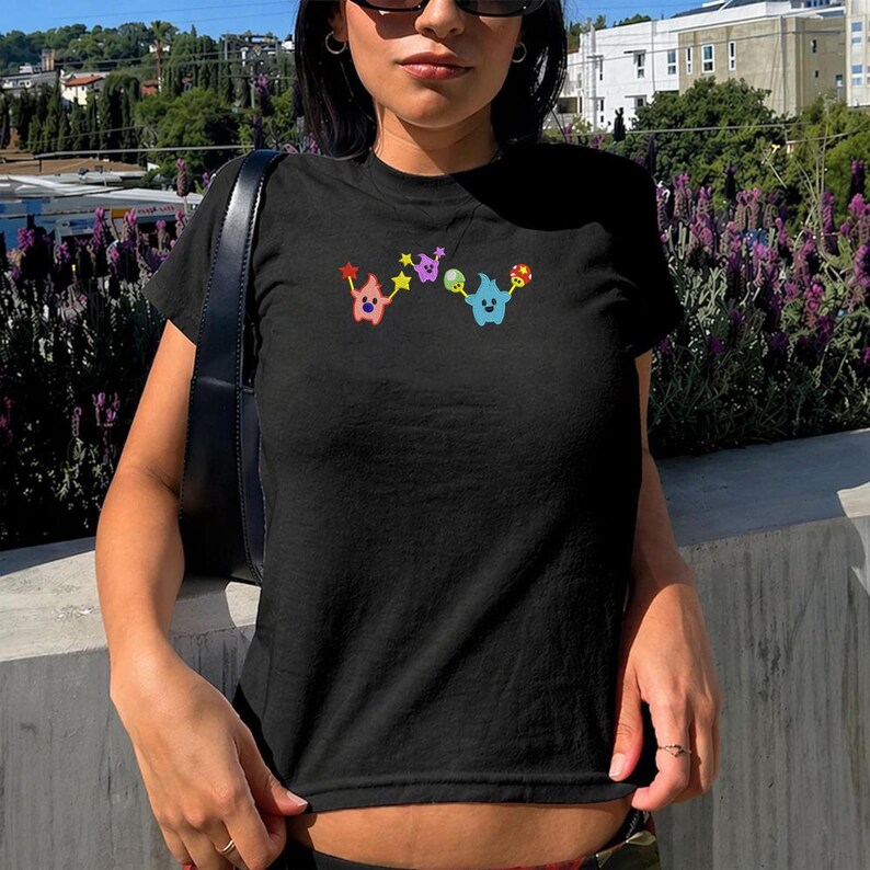 May include: Black crew neck t-shirt with colorful cartoon characters embroidered on the front. The design features two whimsical figures holding stars, musical notes, and a mushroom. The shirt is made of a soft material.