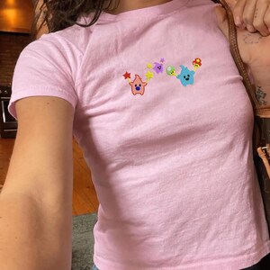 May include: A light pink t-shirt with a crew neck and short sleeves. The shirt features an embroidered design of colorful cartoon characters. The characters include a red star, a purple star, a green mushroom, and a blue blob.