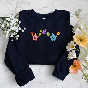 May include: Navy blue sweatshirt with colorful embroidered cartoon characters. The design features three whimsical creatures holding stars, mushrooms, and other shapes. The sweatshirt is made of a soft material and has a crew neck.