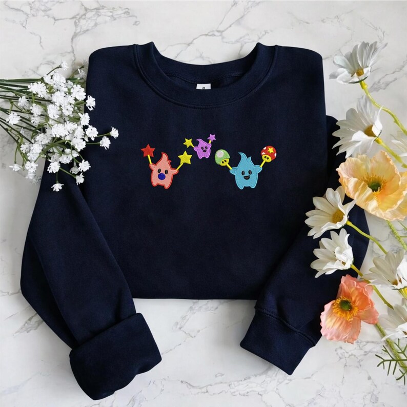 May include: Navy blue sweatshirt with colorful embroidered cartoon characters. The design features three whimsical creatures holding stars, mushrooms, and other shapes. The sweatshirt is made of a soft material and has a crew neck.