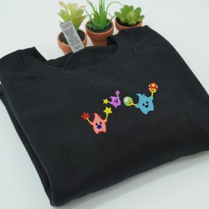 May include: Black crewneck sweatshirt with colorful embroidered cartoon characters. The design features a pink character with red star wands, a purple character with yellow stars, and a blue character with a mushroom and a red and yellow ball.
