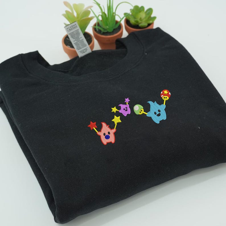May include: Black crewneck sweatshirt with colorful embroidered cartoon characters. The design features a pink character with red star wands, a purple character with yellow stars, and a blue character with a mushroom and a red and yellow ball.