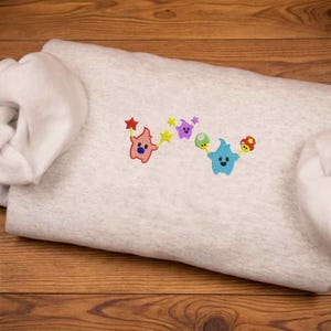 May include: A light gray sweatshirt with colorful embroidered star-shaped characters. The characters are in shades of pink, purple, and blue, each holding a star or a mushroom. The sweatshirt is folded on a wooden surface.
