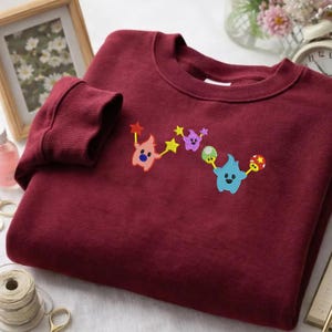 May include: A maroon sweatshirt with embroidered cartoon characters in pink, purple, and blue, holding stars and mushroom-like objects. The design is connected by yellow stars. The sweatshirt has ribbed cuffs and a crew neck.