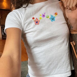 May include: White t-shirt with a crew neck and short sleeves. The shirt features a colorful graphic of cartoon-like characters, including a pink star, purple star, green mushroom, and blue ghost-like figure. The design is centered on the front of the shirt.