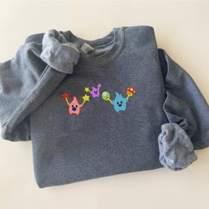 May include: A dark gray sweatshirt with a colorful embroidered design. The design features two cartoon characters holding stars and other shapes in red, purple, green, and blue. The sweatshirt is folded on a white surface.
