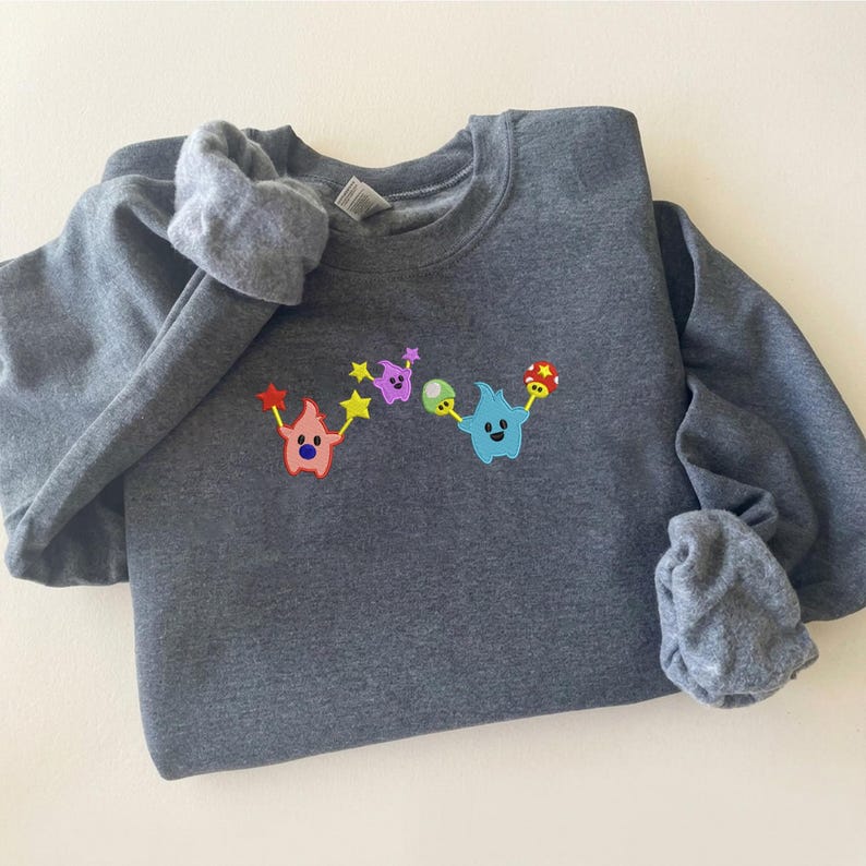 May include: A dark gray sweatshirt with a colorful embroidered design. The design features two cartoon characters holding stars and other shapes in red, purple, green, and blue. The sweatshirt is folded on a white surface.