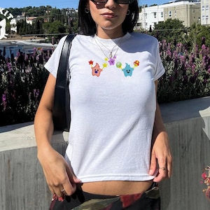 May include: White t-shirt with a crew neck featuring colorful cartoon characters. The design includes a pink star-shaped character, a purple star, a green and blue character, and a mushroom. The shirt is made of a soft, comfortable material.