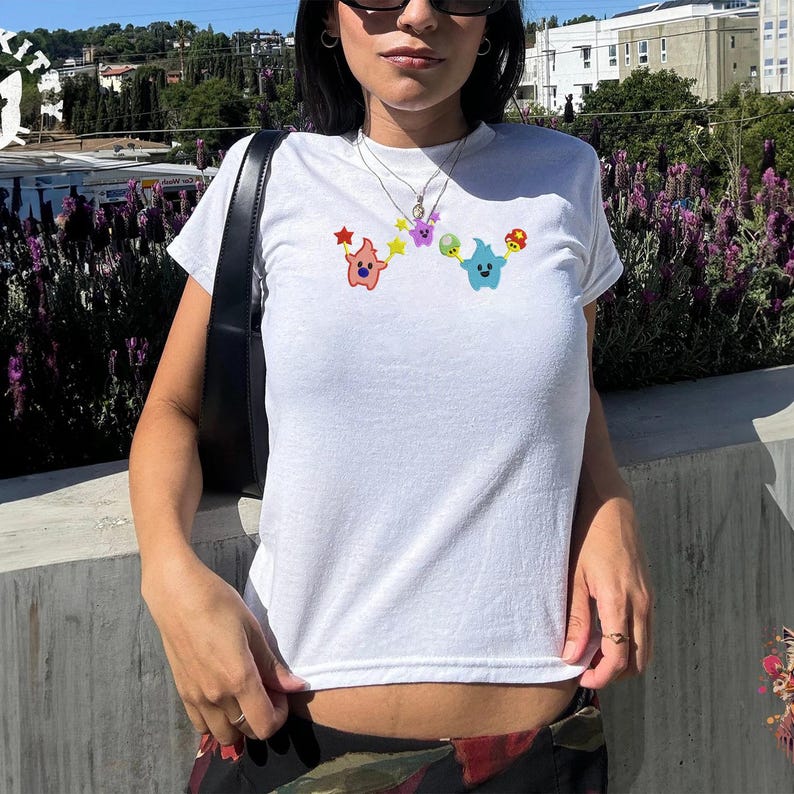 May include: White t-shirt with a crew neck featuring colorful cartoon characters. The design includes a pink star-shaped character, a purple star, a green and blue character, and a mushroom. The shirt is made of a soft, comfortable material.