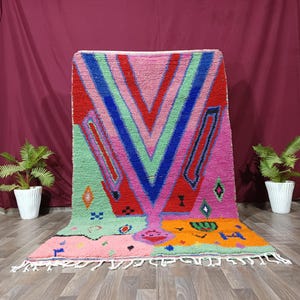 May include: A rectangular area rug featuring a vibrant geometric pattern with large V-shaped stripes in red, blue, and light green against a pink background. The lower section includes orange and pink blocks with small black, blue, and green symbols and fringe.