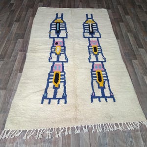 May include: A rectangular off-white area rug featuring two symmetrical vertical columns of geometric patterns in blue, yellow, pink, and black. The rug has a textured surface and is finished with a fringe of white tassels along the bottom edge.