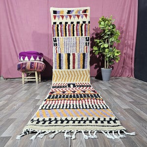 May include: A long, rectangular runner rug featuring a cream-colored base with a geometric pattern in black, yellow, purple, and red. The design includes horizontal stripes, chevron motifs, and block shapes, finished with white fringe at the ends.