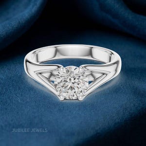 May include: A silver-toned ring featuring a single round-cut clear gemstone set in a split-shank band. The metal has a polished, reflective finish. The text JUBILEE JEWELS is printed in the bottom left corner of the frame.