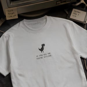 Op de afbeelding: Wit T-shirt met ronde hals met een gepixeld dinosaurusontwerp en de tekst "IF YOU SEE ME. YOU'RE OFFLINE." Een vintage computermonitor en diskette zijn op de achtergrond te zien.
