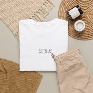 May include: A white short-sleeved t-shirt featuring the black printed text "WRITING MY OWN STORY" centered on the chest, accompanied by a small graphic of a hand holding a pen positioned directly below the final word of the text.
