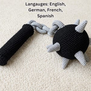 May include: A crocheted flail toy featuring a black cylindrical handle connected by a silver crocheted chain to a black spherical head. The head is adorned with several silver crocheted spikes protruding from its surface. The text reads Langauges: English, German, French, Spanish.