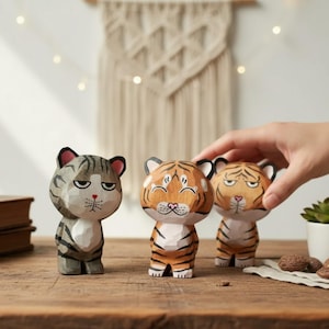 Puede incluir: Tres figuras de tigre de madera con detalles pintados a mano. Los tigres tienen rayas naranjas y negras, grises y negras, y una combinación de colores neutros. Las figuras están sobre una superficie de madera.