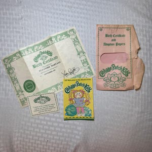 May include: A collection of Cabbage Patch Kids memorabilia, including a birth certificate, adoption papers, and a small card with text. The birth certificate is green and white, and the adoption papers are in a pink envelope. A small yellow card features a Cabbage Patch Kid.