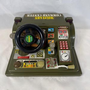 May include: An olive green plastic military command center toy featuring a black steering wheel with a center shot button, a gear shift lever, a numeric keypad, a power switch, and various printed graphics including the text MILITARY COMMAND CENTER.