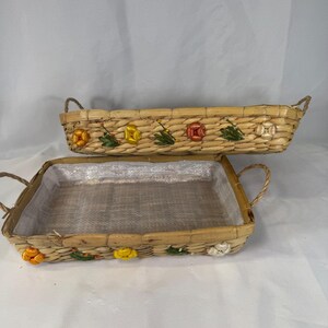 May include: Two rectangular woven baskets with handles, featuring a light tan natural fiber construction. The exterior sides are decorated with small, colorful fabric flowers in yellow, orange, and white, accented by green fabric leaves.