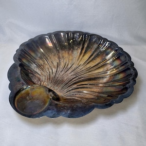 May include: A silver-plated shell-shaped dish featuring a scalloped edge and a deep, fluted interior. The metal surface displays significant natural tarnish with dark, iridescent patches of blue, gold, and brown oxidation throughout the bowl.