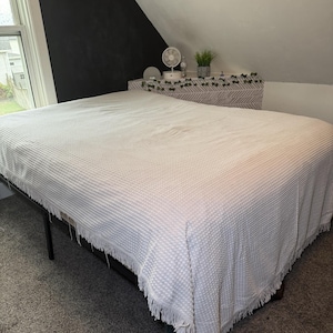 May include: A white textured bedspread with a fringe trim covers a bed frame. The bed is in a room with a window, a black accent wall, and a shelf with decorative items. The bedspread is made of a soft, woven material.