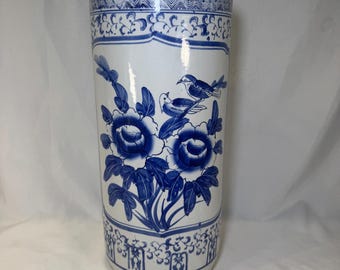 May include: A cylindrical ceramic umbrella stand featuring a white base with blue hand-painted designs. The pattern includes two birds perched on floral branches, surrounded by decorative geometric borders at the top and bottom edges.