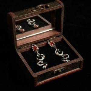 May include: A pair of snake-shaped earrings with red gemstone accents, displayed inside a small wooden jewelry box with a mirrored lid. The earrings feature a white and black enamel design with red details.