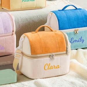 Könnte beinhalten: Vier farbenfrohe, zweistöckige Lunchpakete mit aufgestickten Namen auf der Vorderseite. Jede Tasche hat einen oberen Griff und einen Schultergurt. Farben sind Türkis, Orange, Gelb, Blau, Rosa und Weiß. Die gestickten Namen sind Kiara, Alesia, Emily und Clara.