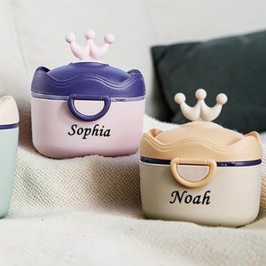 May include: Three personalized baby formula containers in pastel colors: mint green, pink, and beige. Each container has a crown-shaped handle and a name printed on the front: Mateo, Sophia, and Noah. The containers are on a white surface.