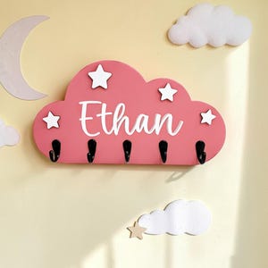 May include: A pink cloud-shaped wall hook rack featuring the name Ethan in white script lettering. The rack is decorated with four white star cutouts and includes five black metal hooks attached to the bottom edge for hanging items.