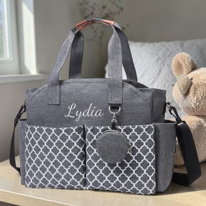 May include: A gray diaper bag with a white quatrefoil pattern on the front pockets. The bag has a shoulder strap and top handles. The name "Lydia" is embroidered on the front. A small, round, matching accessory is attached.
