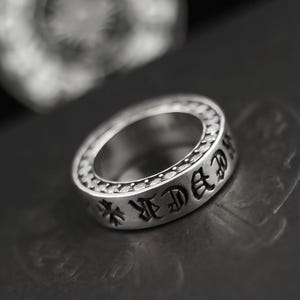 Può includere: Un anello d'argento con scritte incise e disegni a forma di croce. L'anello ha una superficie dettagliata e strutturata e una fascia larga. L'anello è esposto su una superficie scura, evidenziando la sua lucentezza metallica.