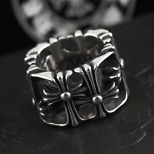 May include: A silver-toned ring with a square design. The ring features a repeating cross motif on the sides and top. The ring has a polished finish and a bold, geometric style.