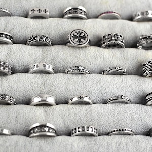Può includere: Una collezione di anelli d'argento esposti su una superficie grigia. Gli anelli presentano vari design, tra cui croci, motivi floreali e forme geometriche. Alcuni anelli hanno pietre preziose incastonate.