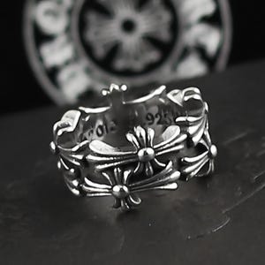 Puede incluir: Un anillo plateado con un diseño de cruz floral. El anillo tiene un patrón detallado de cruces y elementos florales. El anillo está marcado con "925" y otro texto. El anillo está sobre una superficie oscura.