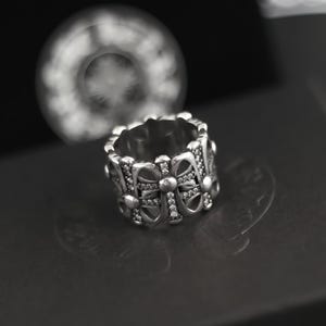 May include: A silver ring with a repeating cross design, embellished with small, sparkling stones. The ring has a detailed, textured appearance and is set against a dark background, highlighting its intricate craftsmanship.
