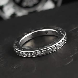 Può includere: Un anello d'argento con una fascia di piccole pietre trasparenti. L'anello ha un disegno a croce sulla parte superiore e testo sul bordo. L'anello è su una superficie scura.