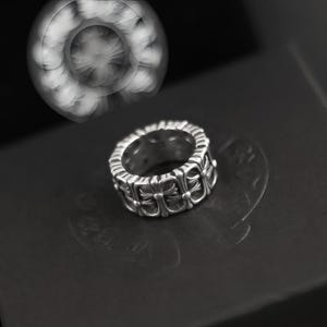 May include: A silver ring with a repeating cross design. The ring is made of a shiny metal and has a detailed pattern of crosses around its circumference. The ring is shown on a dark surface.