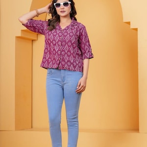 Pode incluir: Uma pessoa vestindo uma camisa de manga curta com botões com um padrão geométrico em tons de roxo e branco, combinada com jeans azul claro. Eles também estão usando óculos de sol brancos.