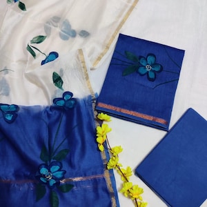May include: A white and blue floral print salwar kameez set. The set includes a sheer white dupatta with blue flower designs, a blue top with gold trim, and a solid blue bottom. Yellow flowers are placed near the set.