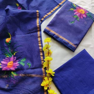 Pode incluir: Um conjunto de peças de tecido azul índigo, incluindo um lenço e dois painéis retangulares. Um painel apresenta um grande desenho floral em rosa, laranja e verde. A borda dourada orla o tecido. Um raminho de flores amarelas adorna a exibição.