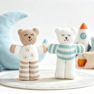 Könnte beinhalten: Zwei gestrickte Bärenpuppen stehen nebeneinander. Der linke Bär ist beige-weiß mit Blumenstickerei auf der Brust und gestreiften Beinen. Der rechte Bär ist weiß mit türkisfarbenen Querstreifen am Oberkörper. Beide haben geschlossene Augen.