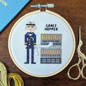 May include: Cross-stitch embroidery in a wooden hoop depicting Grace Hopper in a navy uniform. The text "GRACE HOPPER" is above a computer. Gold scissors and thread are in the background.