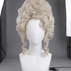 May include: A light blonde wig with voluminous curls and a high, rounded crown, displayed on a white mannequin head. The wig features loose, cascading curls framing the face and neck, creating a classic, historical look.