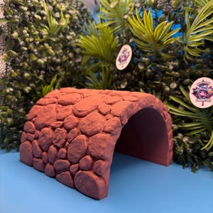 May include: A terracotta-coloured arched tunnel hideout featuring a textured exterior surface that mimics the appearance of stacked stones. The structure has a hollow interior and a wide opening, designed for small animal habitats or enclosures.