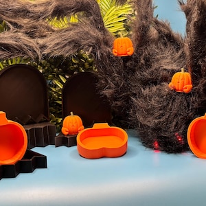 May include: A collection of bright orange pumpkin-shaped containers and black tombstone-shaped display stands. The set includes three small orange pumpkin figurines with carved faces, arranged among the larger containers and a large fuzzy black spider.