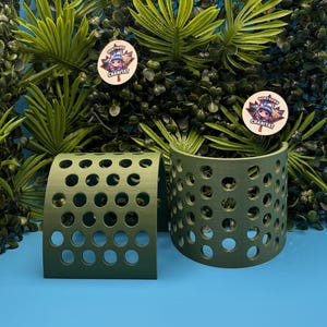 May include: Two olive green, curved, semi-cylindrical structures featuring a repeating pattern of circular cutouts. Each piece has a smooth, matte finish and a hollow interior. Two circular stickers with the text Northern Crabitat are visible.